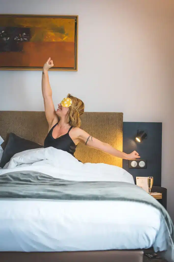 Femme s'étirant au réveil dans le lit double d'une chambre du Masq Hôtel, masque de nuit sur les yeux