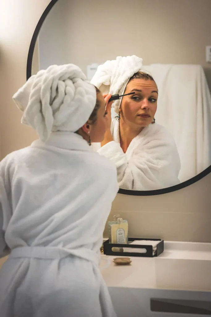 Cliente en peignoir se maquillant devant le miroir rond d'une salle de bain du Masq Hôtel