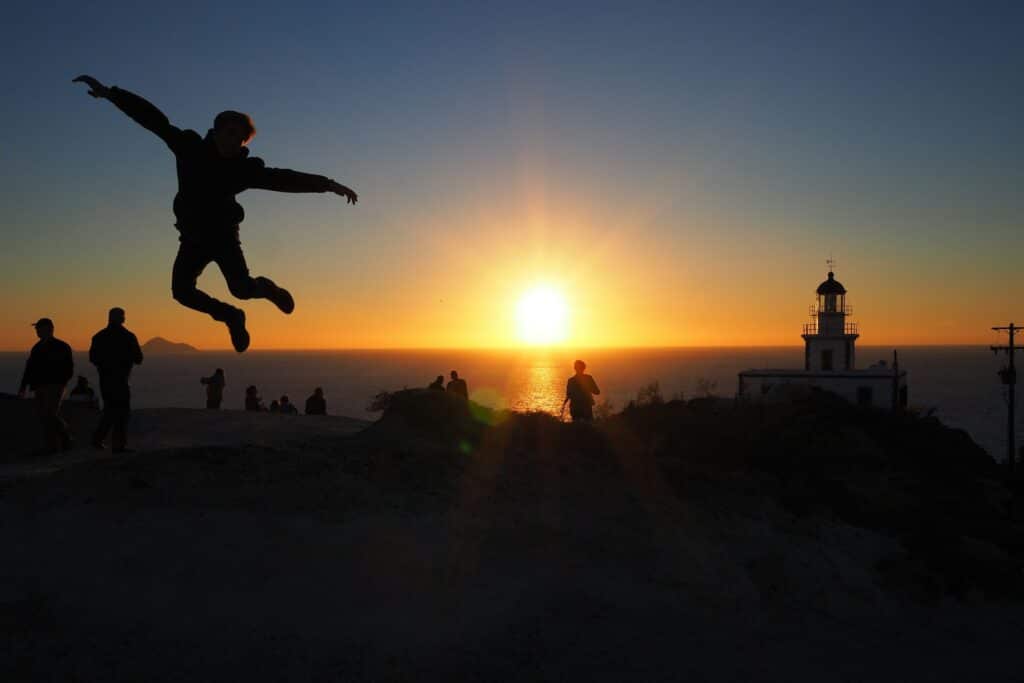 Silhouette d'un homme sautant devant le coucher de soleil au phare d'Akrotiri à Santorin avec visiteurs admirant le spectacle