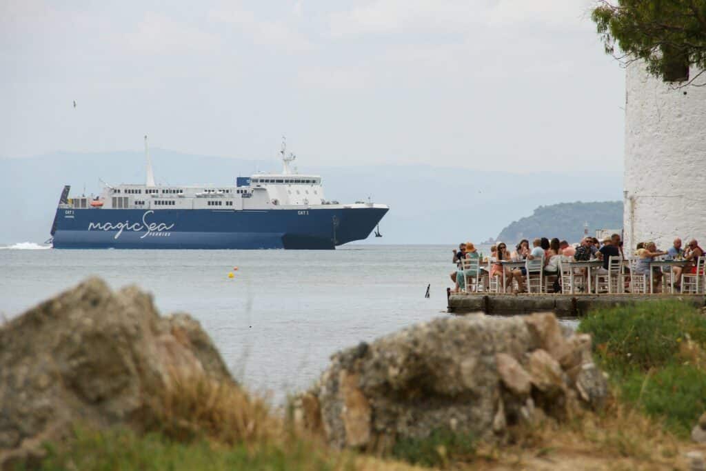 Ferry MagicSea passant devant une taverne grecque où des touristes déjeunent au bord de l'eau