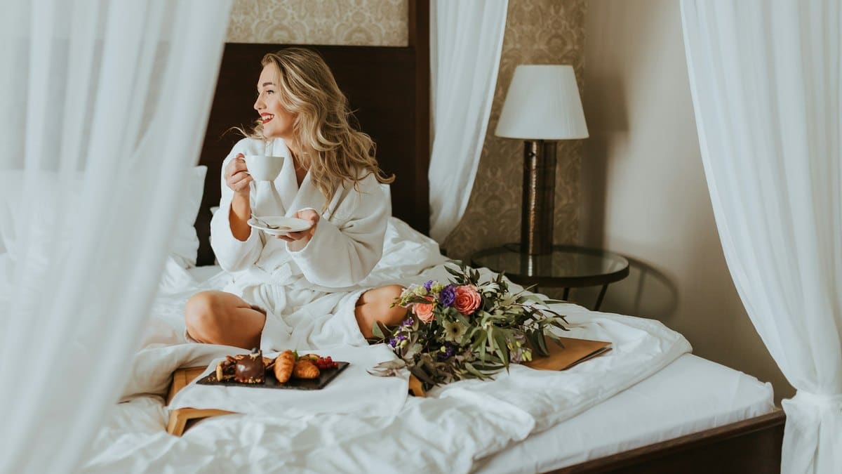 Femme en peignoir buvant un café sur un lit à baldaquin, plateau de petit-déjeuner et bouquet de fleurs