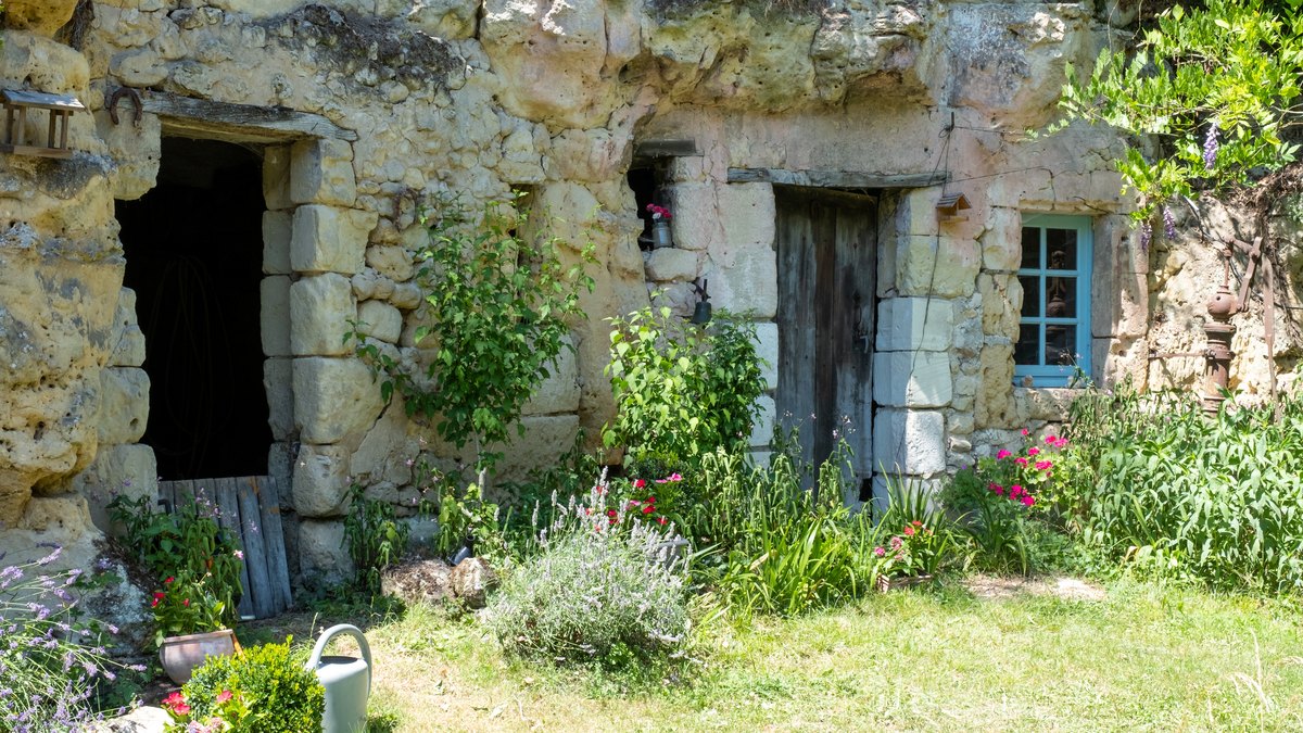 Façade d'une maison troglodytique creusée dans le tuffeau, avec porte en bois, fenêtre bleue et jardin fleuri