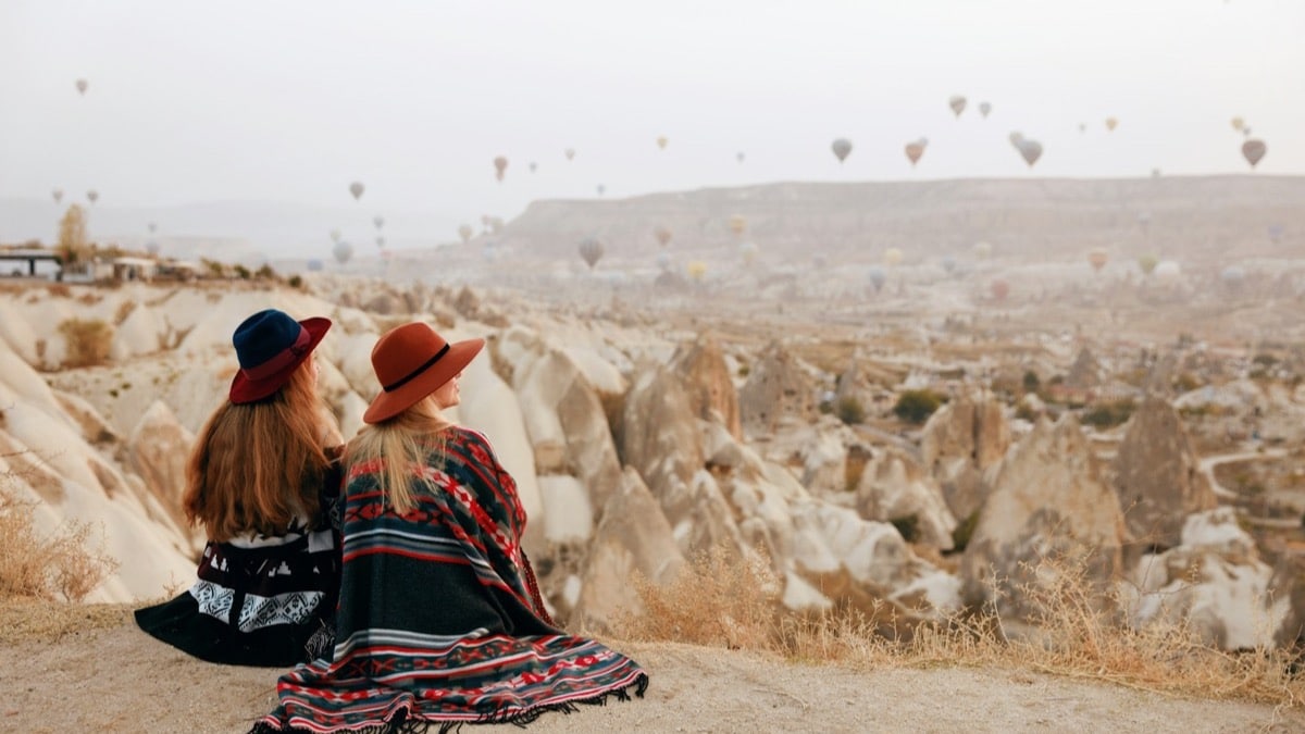 Deux voyageuses en chapeau assises face aux vallées de Cappadoce, montgolfières dans le ciel au lever du jour