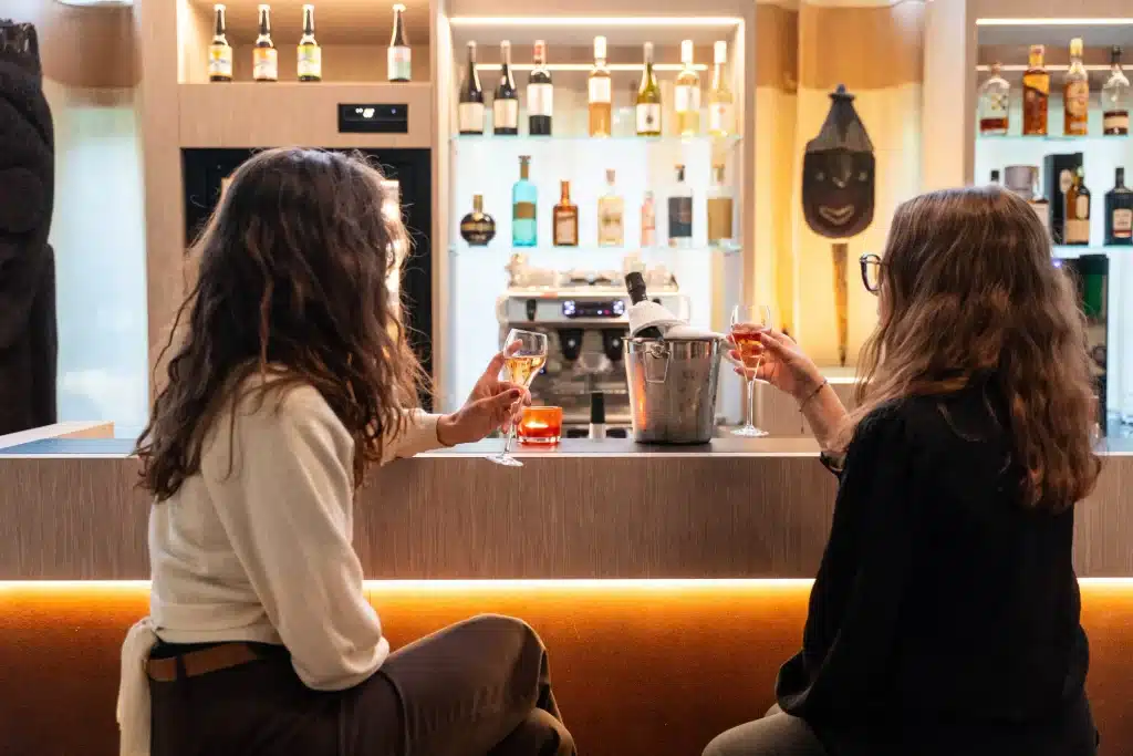 Deux clientes trinquant au champagne au comptoir du bar L'Ouvrage à Cornes, devant des étagères de spiritueux et un masque africain