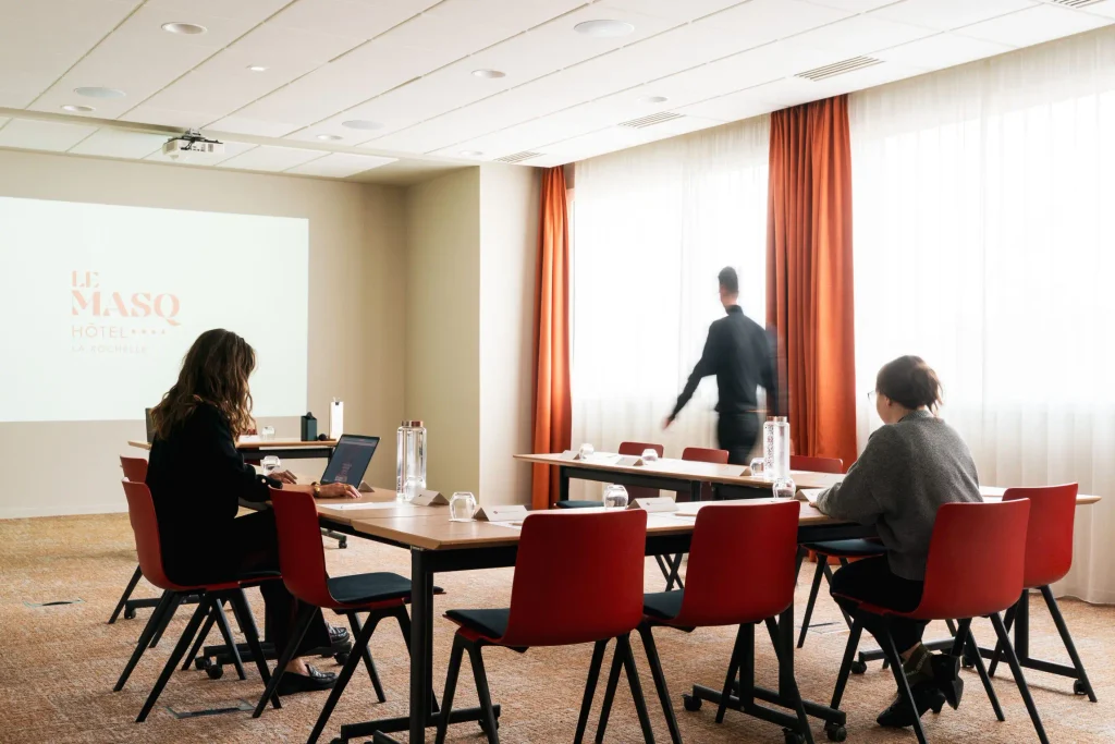 Salle de séminaire lumineuse du Masq Hôtel avec chaises rouges, vidéoprojecteur et écran affichant le logo de l'hôtel
