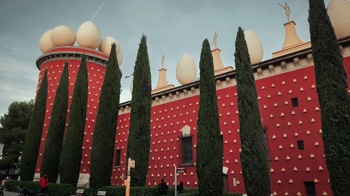 Façade rouge de la tour Galatea du Théâtre-Musée Dalí à Figueres, couronnée d'œufs géants et ponctuée de pains dorés, devant une rangée de cyprès