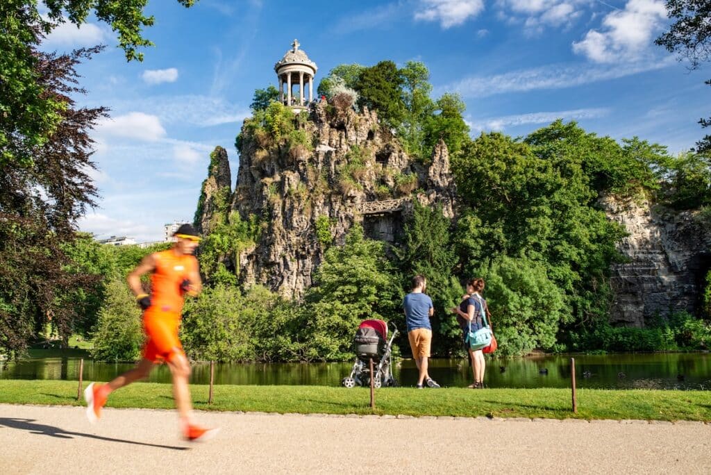 Une famille avec un bébé en poussette admire les falaises du Parc des Buttes-Chaumont tandis qu'un joggeur passe derrière eux.