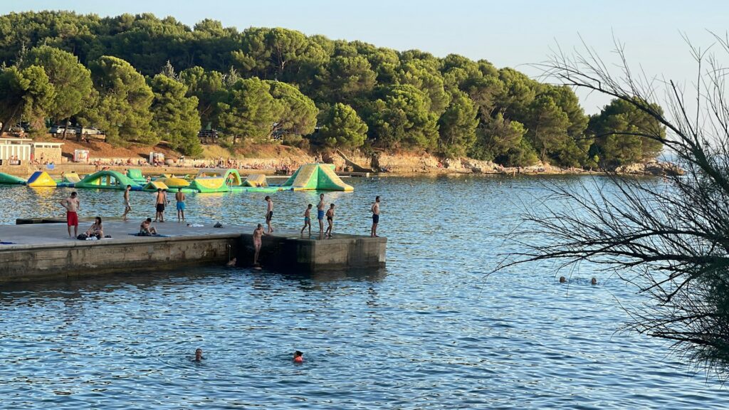 Baigneurs sur un ponton de béton et parc aquatique gonflable devant une côte boisée de pins à Pula