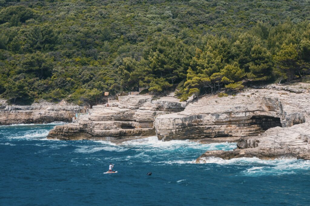 Paddleur agenouillé sur sa planche au pied de falaises calcaires creusées d'une grotte, côte boisée de pins à Pula