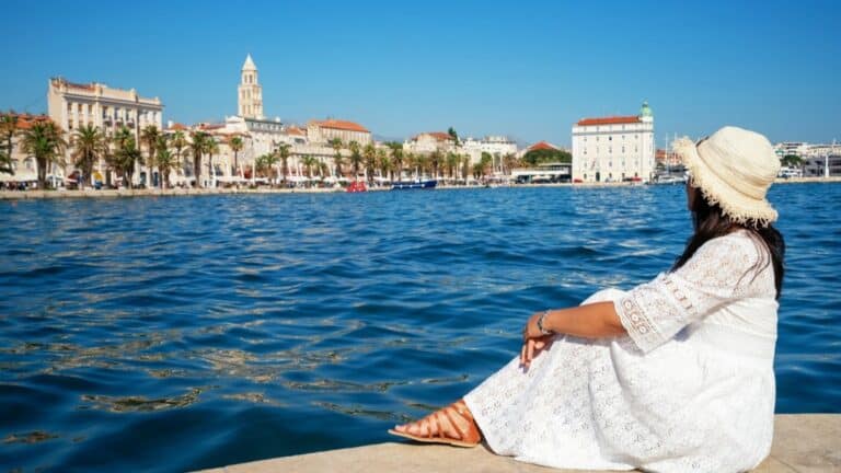 Voyageuse en robe blanche assise au bord de l'eau face à la Riva de Split, aux palmiers et au clocher de Saint-Domnius
