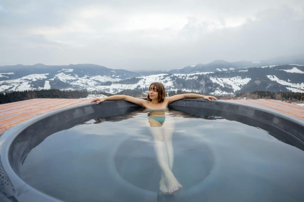 Une femme se prélasse en hiver dans un jacuzzi avec vue sur les montagnes enneigées.