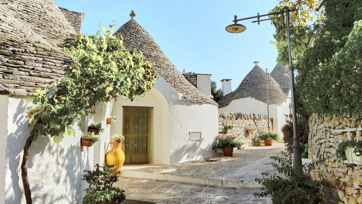 Ruelle pavée bordée de trulli blanchis à la chaux, aux toits coniques en pierre, avec une jarre jaune et des plantes en pot
